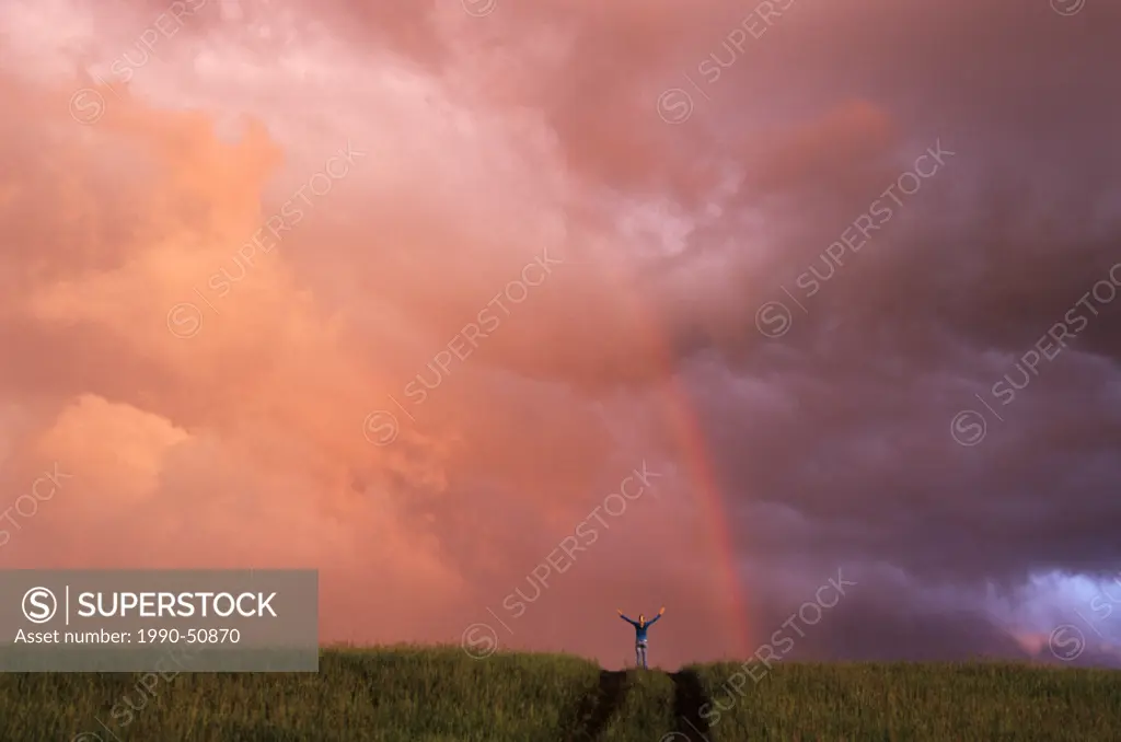 Girl on hill with arms outstretched, in front of rainbow, near Winnipeg, Manitoba, Canada.