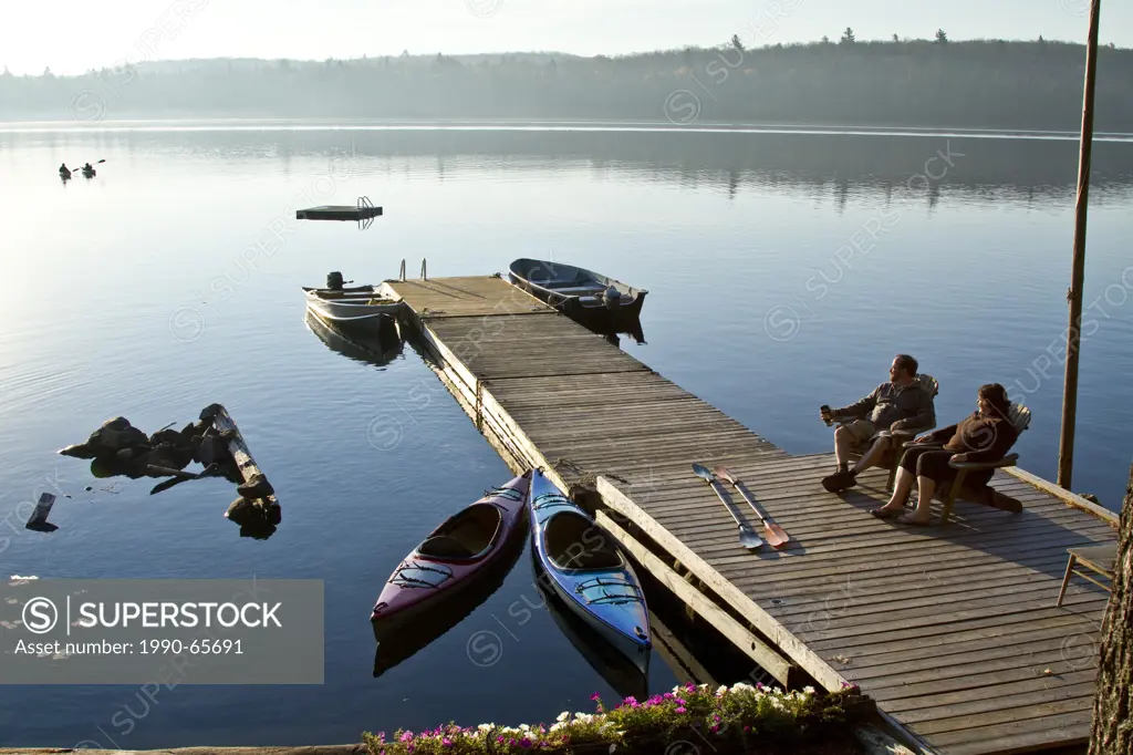 Middle_aged couple enjoy quiet morning on dock near their cottage while thier children kayak on Source Lake, Algonquin Park, Ontario, Canada.