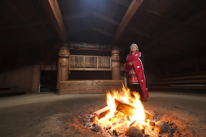 A first nations elder dressed in her traditional regalia stands beside a fire in the bighouse in the village of Oweekeno. Wuikinuxv, Rivers Inlet, Bri...