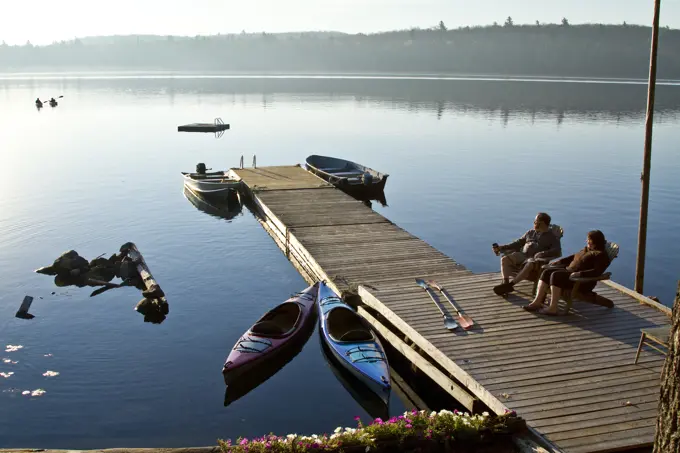 Middle_aged couple enjoy quiet morning on dock near their cottage while thier children kayak on Source Lake, Algonquin Park, Ontario, Canada.