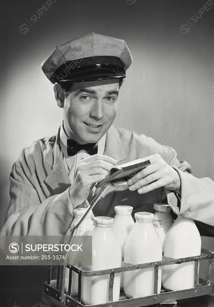 Milkman looking at camera in uniform writing on note pad sitting on case of milk bottles