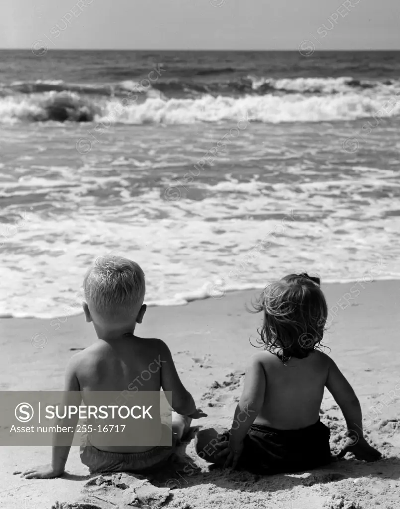 Rear view of pair of children sitting on beach, watching surf