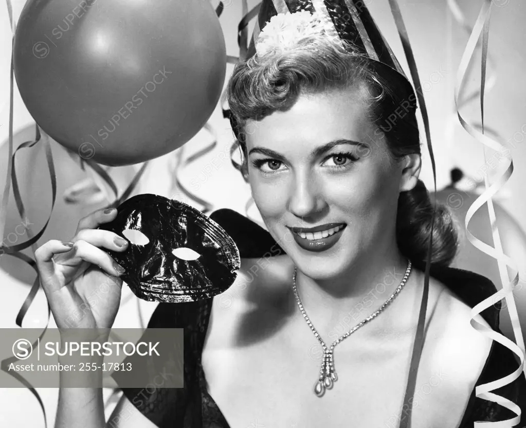 Portrait of beautiful young woman holding carnival mask with balloons and streamers in background