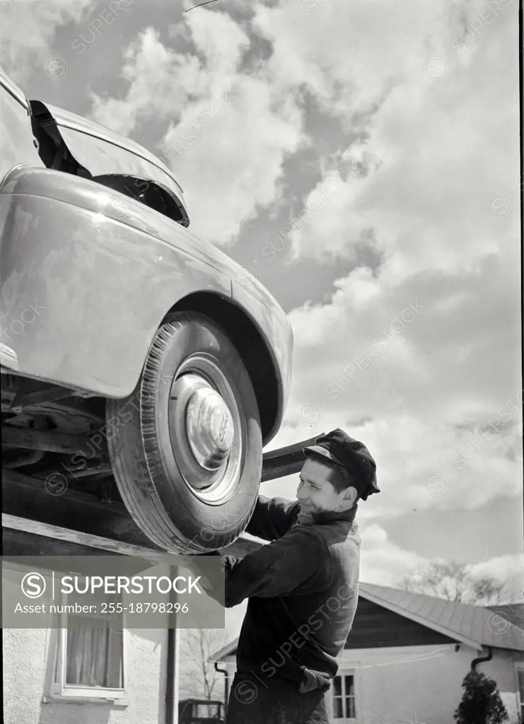 Vintage photograph. Auto mechanic working on car raised up on auto lift outside in Massapequa, Long Island, New York