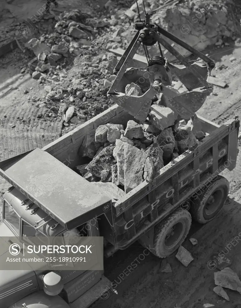 Vintage photograph. Close up of giant claw on the end of a crane unloading large rocks from construction site onto truck bed.