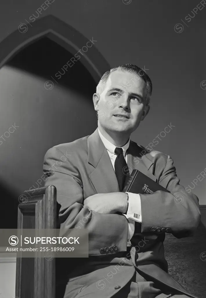 Vintage photograph. Man in suit and tie sitting in church pew looking up with slight smile