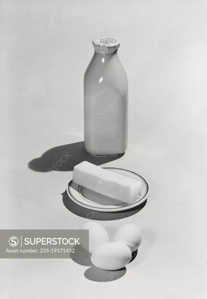 Vintage photograph. Bottle of milk, butter on plate, and three eggs lined up on light background