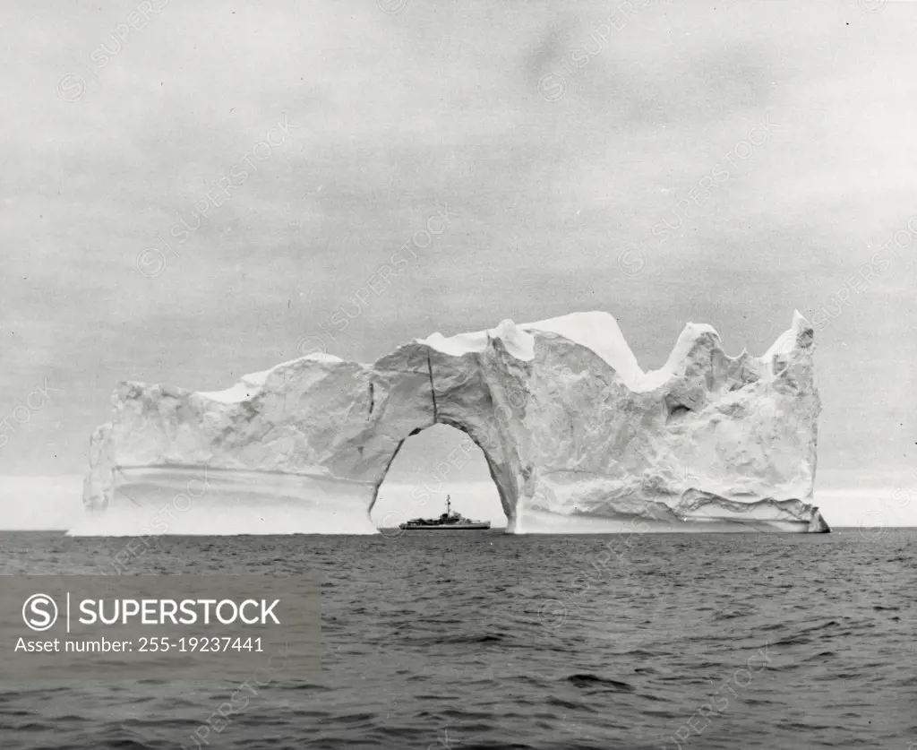 Vintage photograph. An iceberg of tremendous size on its way through the Baffin Bay makes a Frosty frame for the US Coast Guard cutter east wind seen through a natural arch in the berg