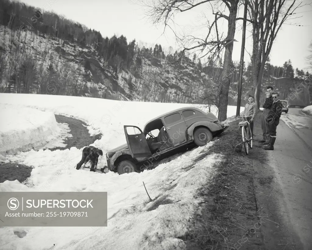 Vintage photograph. Man digging out car in snow filled ditch with onlookers