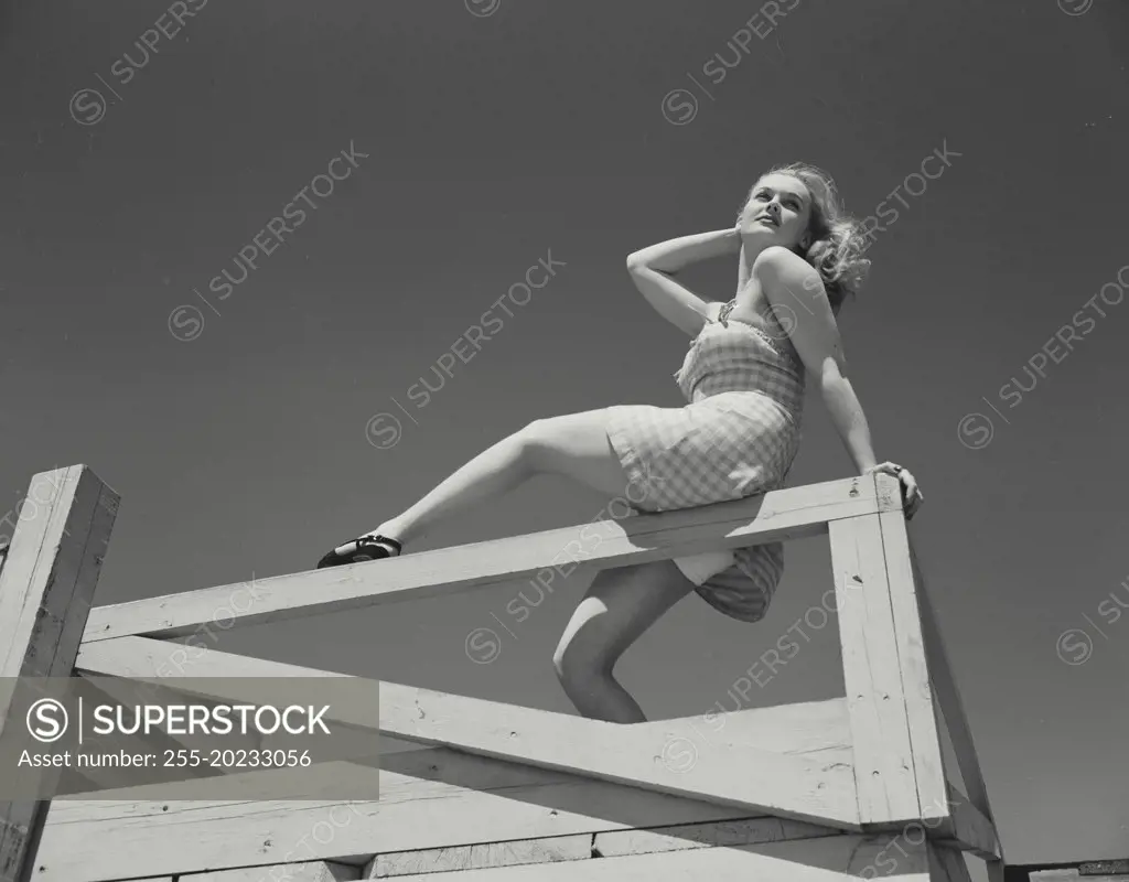 Woman in short dress sitting on railing of deck looking over shoulder