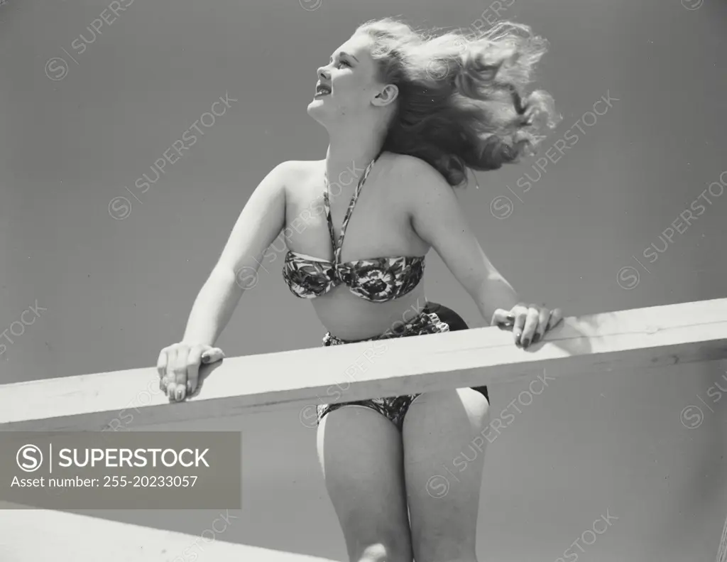 Woman in bikini leaning over railing of deck with hair blowing in wind