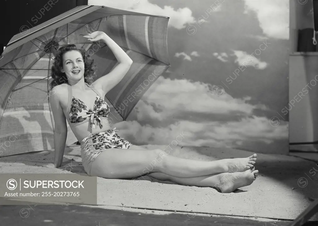 Vintage photograph. Smiling curly haired woman wearing swimsuit lounging with legs outstretched in sand on beach set with large umbrella and sky backdrop with arm raised up