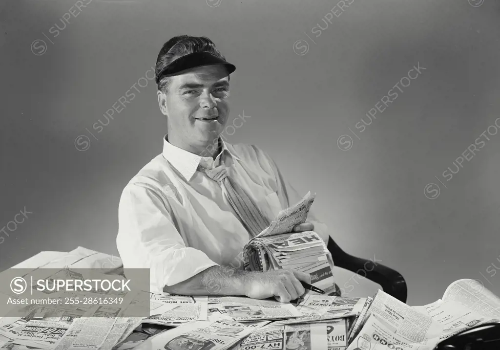 Vintage Photograph. Businessman wearing visor sitting back at office desk covered in newspaper clippings smiling at camera