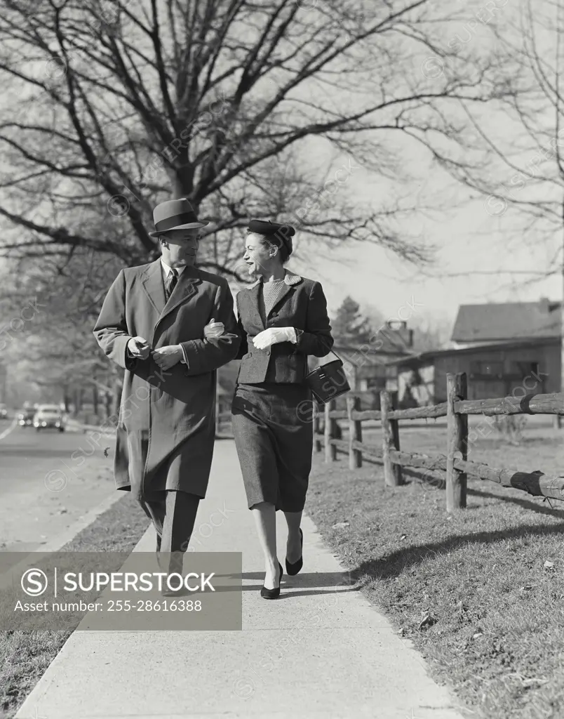 Vintage Photograph. Well dressed man and woman walking arm in arm down  sidewalk with bare tree in background Frame 1 - SuperStock