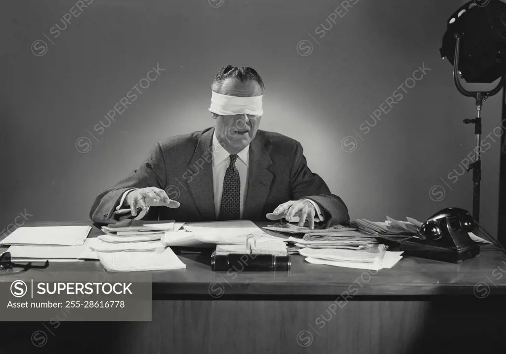 Vintage Photograph. Blindfolded man sitting at desk covered in documents. Frame 1