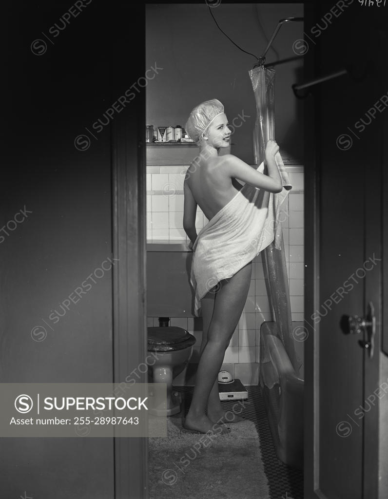 Vintage Photograph. Nude woman in bathroom wearing only shower cap drying  off with towel, viewed through doorway - SuperStock