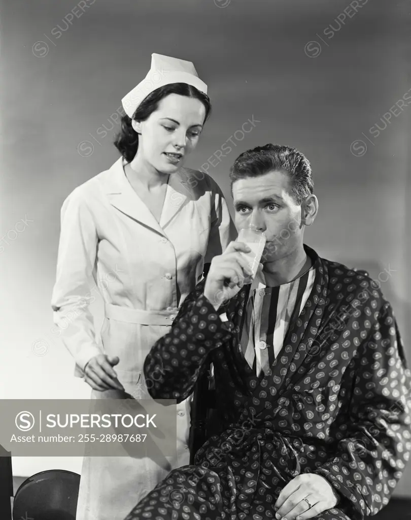 Vintage Photograph. Man wearing silk robe drinking glass of milk with nurse  standing behind him - SuperStock