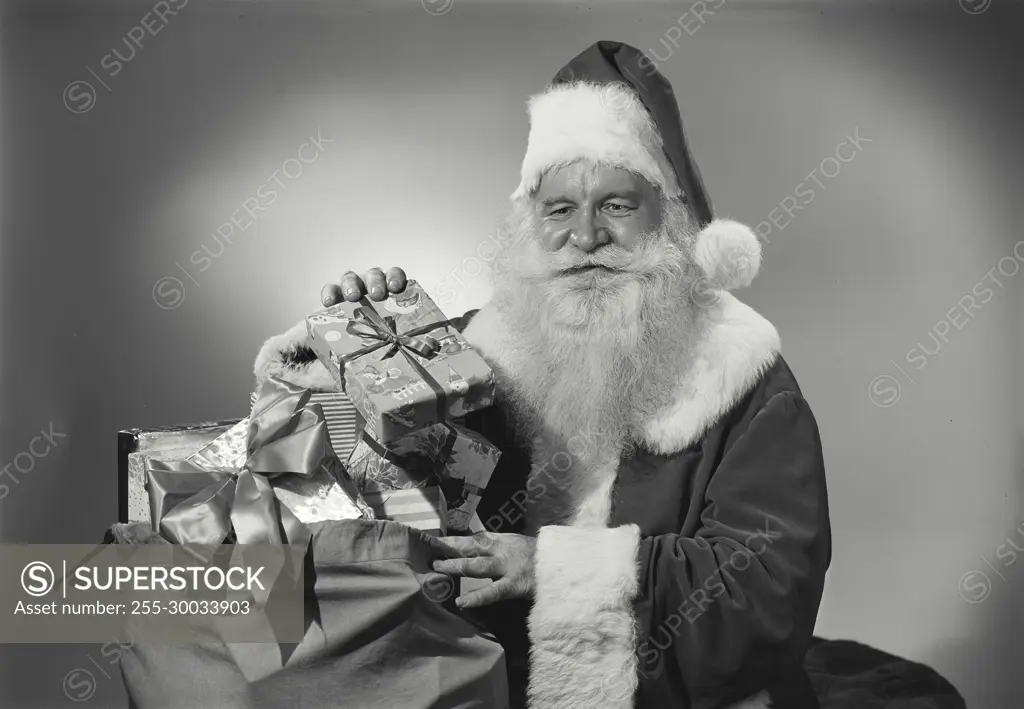 Vintage Photograph. Man in Santa Claus outfit takes gifts out of sack as he smiles.