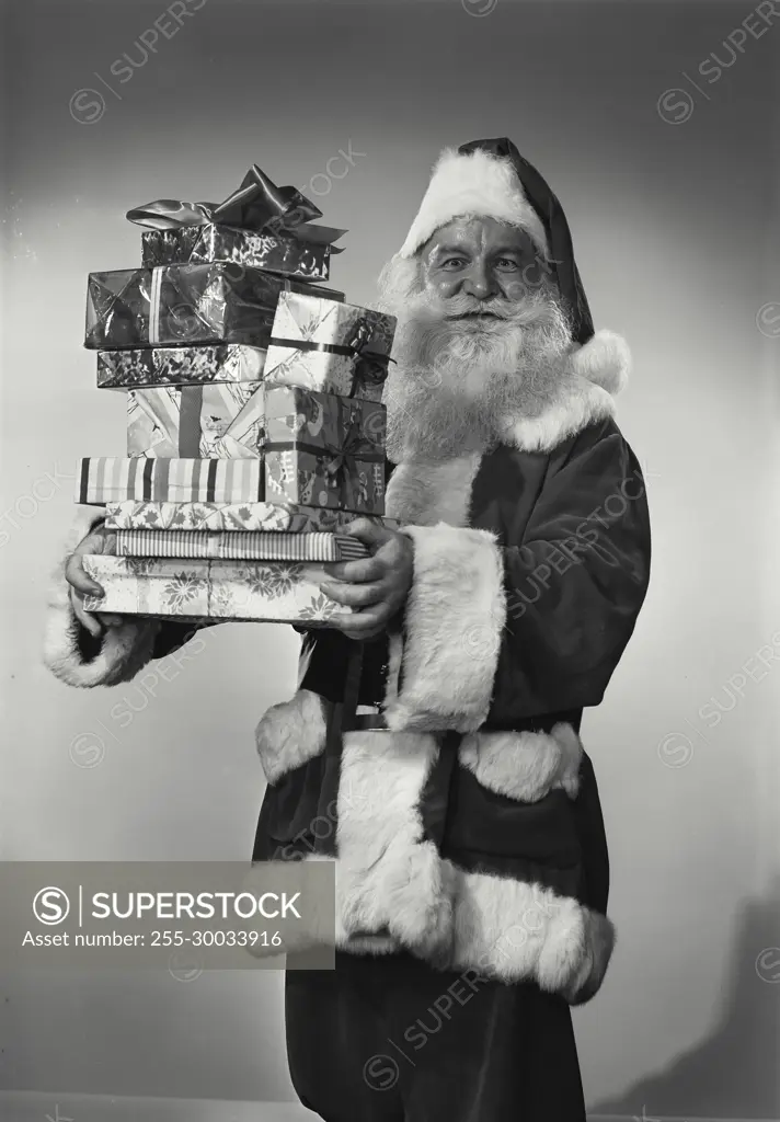 Vintage Photograph. Man in Santa Claus outfit holds up stack of gifts with wide eyes.