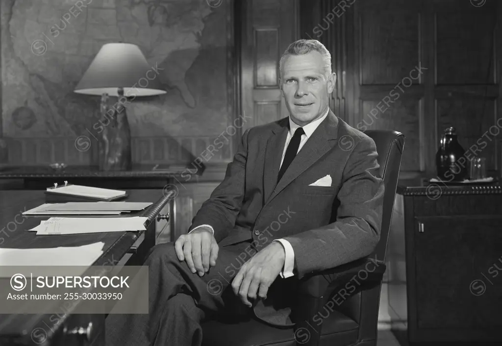 Vintage Photograph. Man sits at desk facing camera with legs crossed.