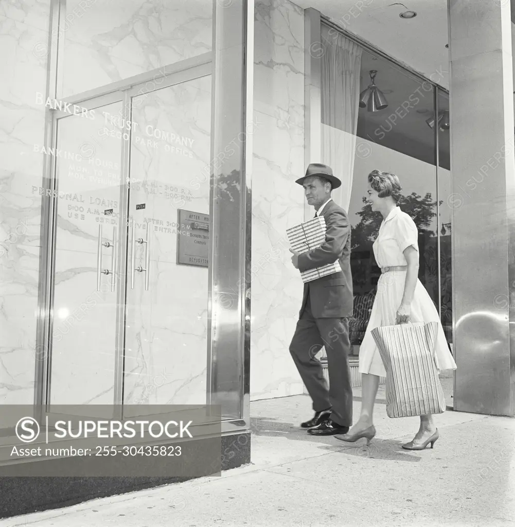 Vintage Photograph. Man and woman walk into front of bank building man carries packages while woman carries a bag.