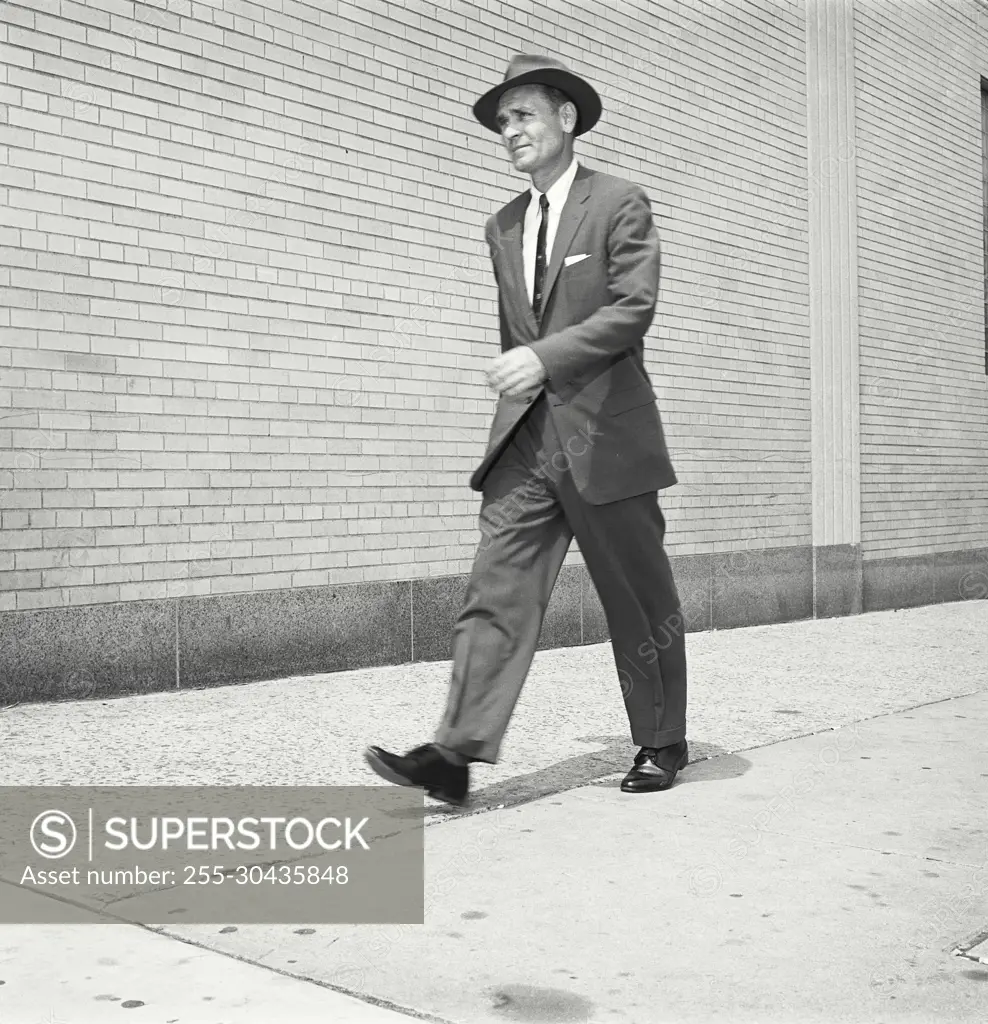 Vintage Photograph. Man walks down city sidewalk wearing suit tie and hat.