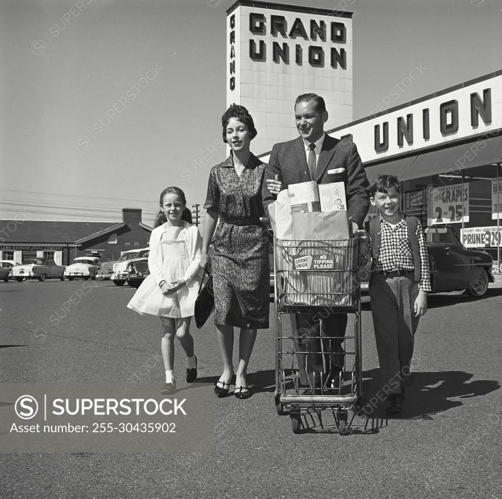 Vintage Photograph. Man pushes family's grocery cart in parking lot as the family leaves the grocery store.