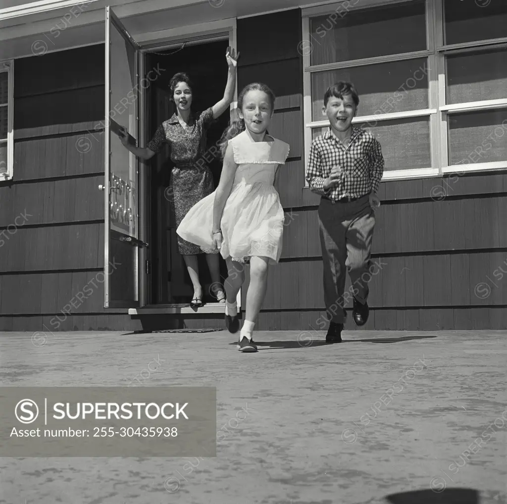 Vintage Photograph. Woman waves as two kids run towards the camera from front of house.