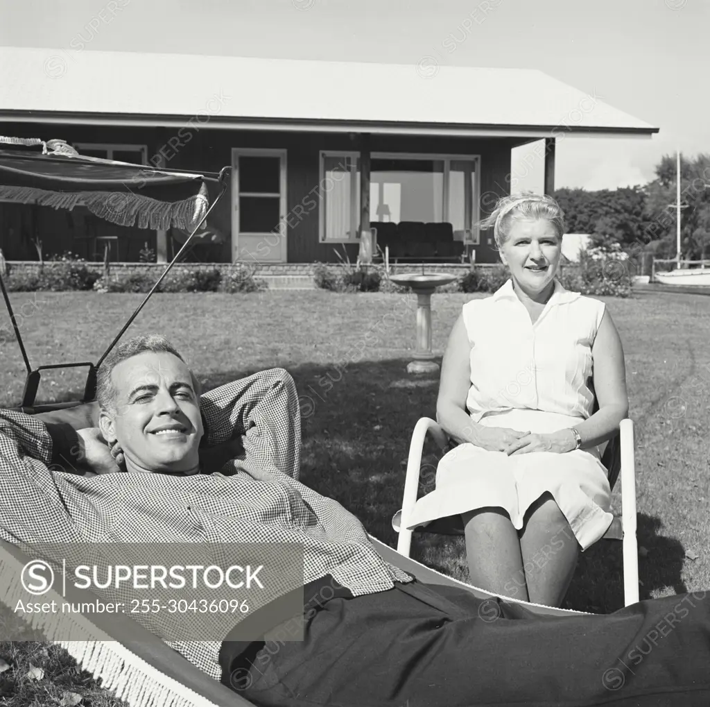 Vintage Photograph. Man relaxes in hammock while woman sits beside him.