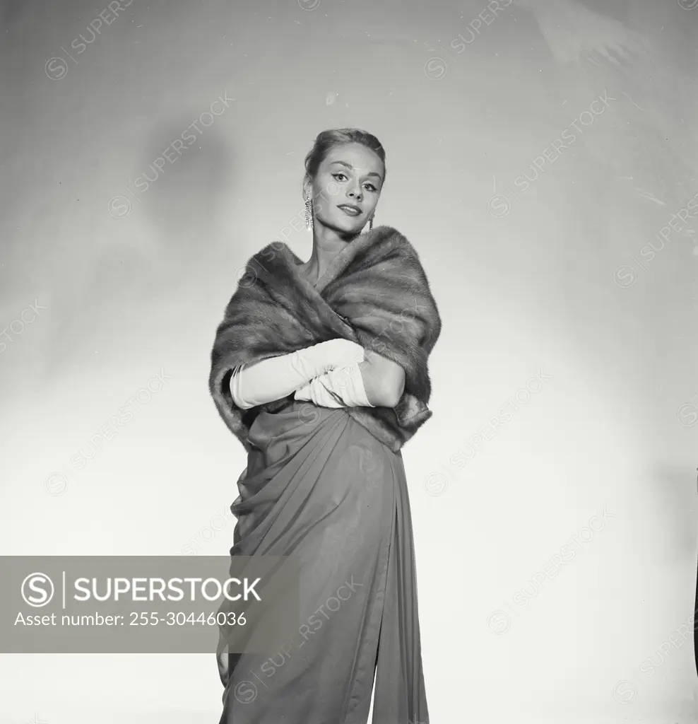 Vintage Photograph. Woman wears formal attire including dress and fur coat as she poses for studio portrait.