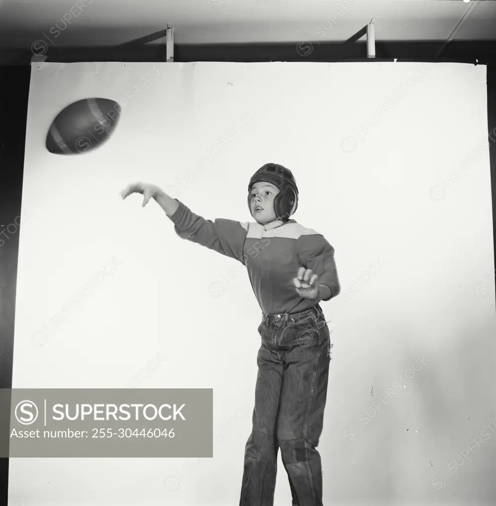 Vintage Photograph. Young boy throws football to the left of frame as he wears jeans and football uniform.