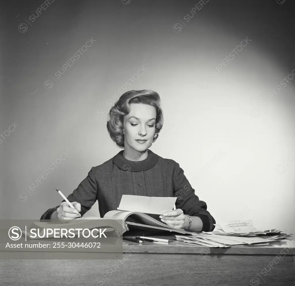 Vintage Photograph. Woman sits behind desk working on paperwork she takes  note of reciept into book. - SuperStock