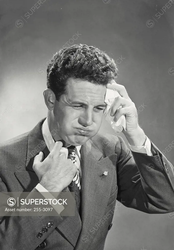 Man with curly dark hair wearing suit and tie holding handkerchief to forehead puffing out cheeks looking uncomfortable