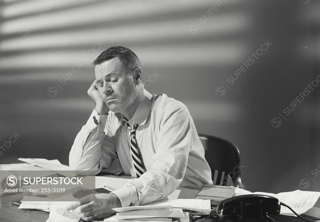 Vintage Photograph. Man in dress shirt and striped tie sitting at desk covered with papers looking exhausted