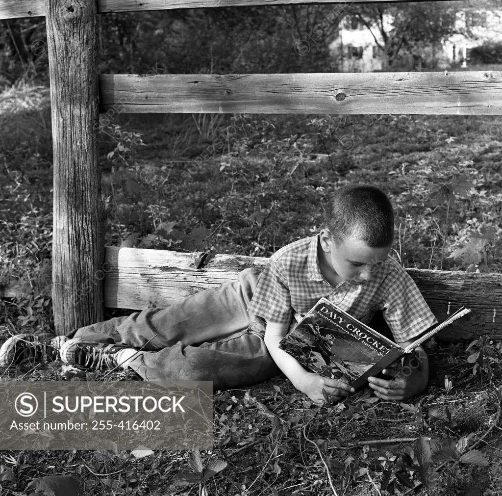 Boy lying on grass by wooden fence and reading book