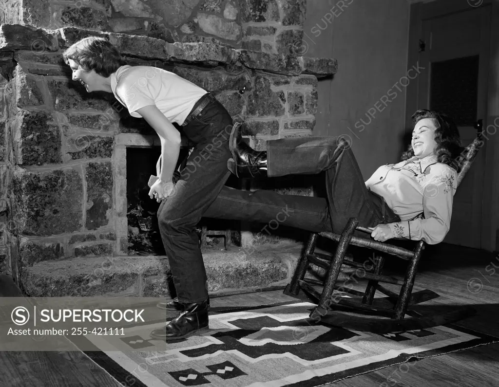 Woman pulling cowboy boot of another's in front of fireplace