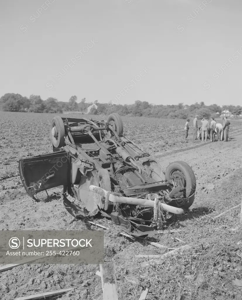 Overturned car in a field, crowd gathered near injured.