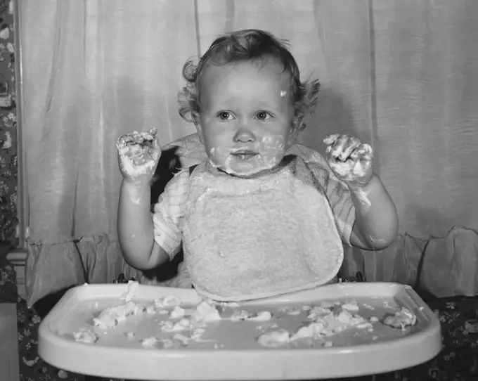 Close-up of a baby sitting in a high chair with food on his face and hands