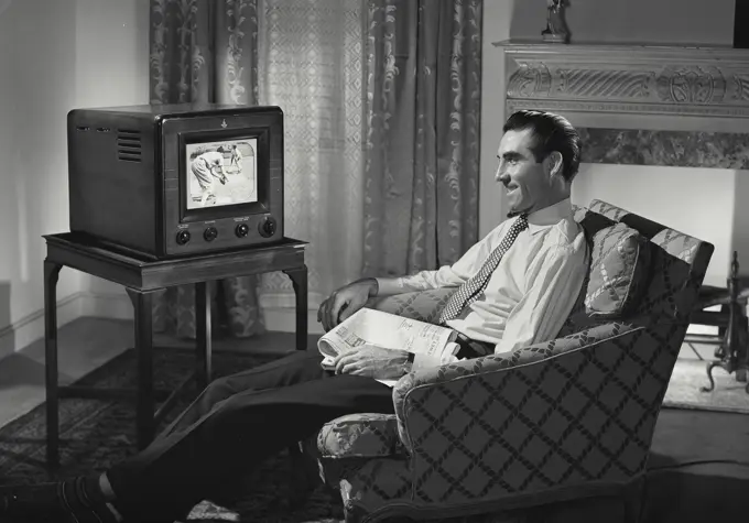 Vintage Photograph. Man relaxing in chair in living room with newspaper in lap watching television with baseball players on screen