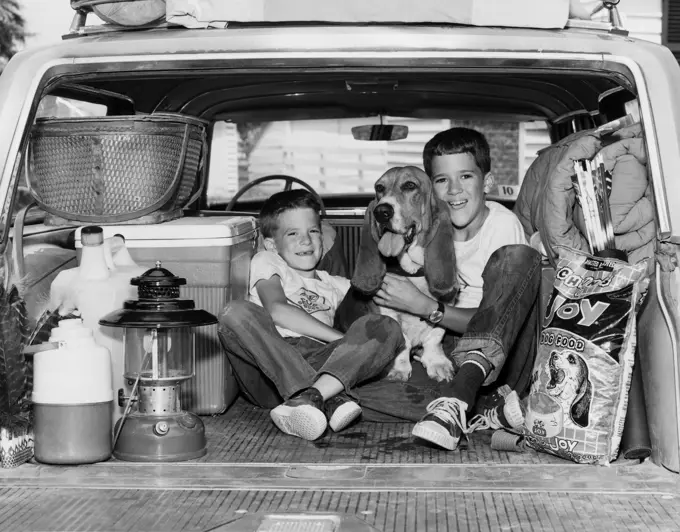 Two boys sitting with their dog in the back of a station wagon