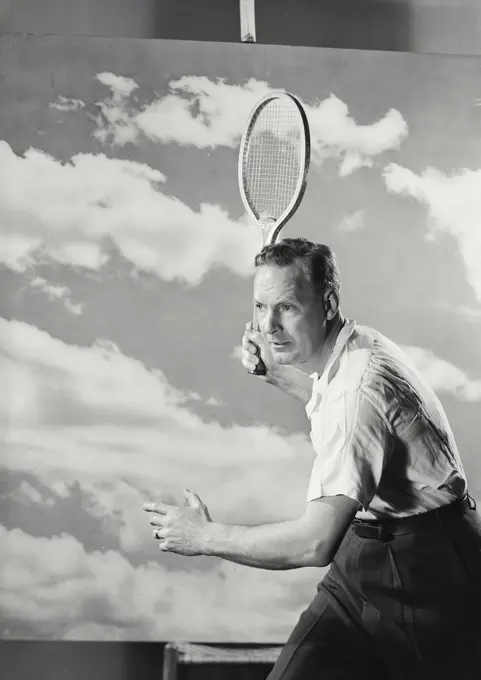 Vintage Photograph. Middle aged man in white shirt and trousers swinging tennis racket in front of cloud background