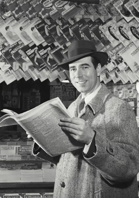 Vintage photograph. Man in coat and hat holding newspaper in front of news stand