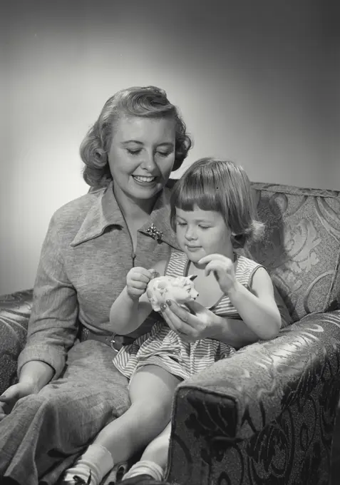 Vintage photograph. Mother and daughter sit together and put coins into piggy bank