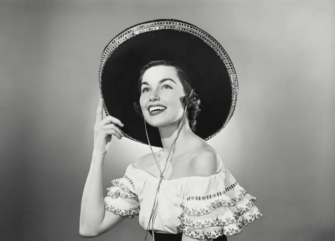 Vintage photograph. Brunette woman wearing traditional Mexican dress and hand raised to black sombrero with silver trim detail