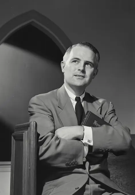 Vintage photograph. Man in suit and tie sitting in church pew looking up with slight smile