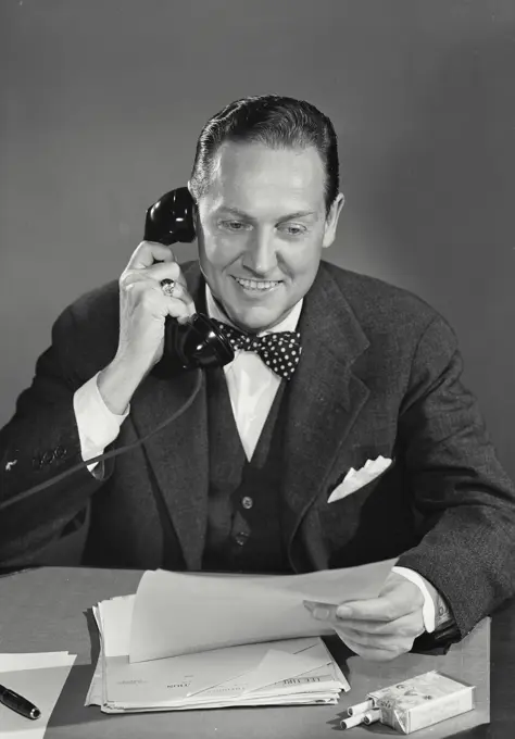 Vintage Photograph. Businessman with bow tie holding phone receiver looking at documents