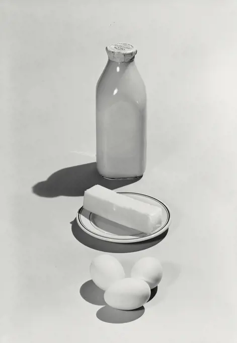 Vintage photograph. Bottle of milk, butter on plate, and three eggs lined up on light background