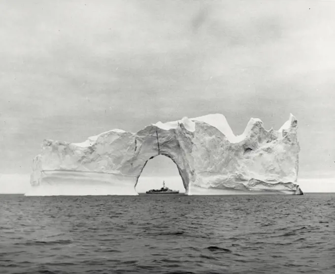 Vintage photograph. An iceberg of tremendous size on its way through the Baffin Bay makes a Frosty frame for the US Coast Guard cutter east wind seen through a natural arch in the berg