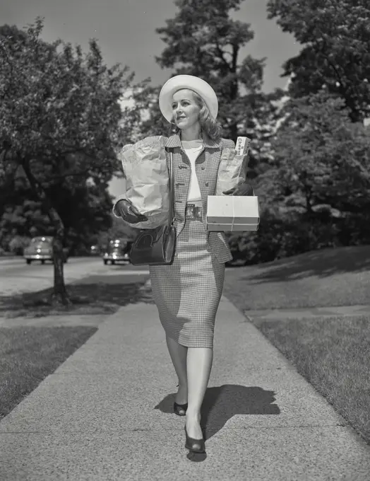 Vintage photograph. Woman in matching skirt and jacket on sidewalk carrying groceries