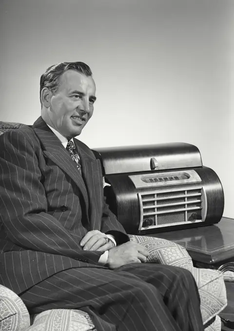 Vintage Photograph. Caucasian man wearing pin striped suit sitting in arm chair next to large radio